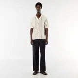 Camisa de manga corta confeccionada en tejido de punto fantasía color beige, con cuello camisero, cierre frontal de botones y detalles de líneas oscuras en zigzag.