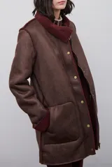 Chaquetón de efecto cuero en color marrón, con interior y detalles en corderito color bordó. Presenta cierre frontal con cremallera y botones a presión, cuello solapa y mangas largas con puños de corderito.