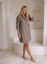 Vestido camisero de lino verde militar, con cuello camisero, botonadura frontal completa con botones de nácar, un bolsillo delantero y mangas largas con lengüeta para ajustar. Diseño de corte recto con dobladillo asimétrico más largo en la parte trasera.