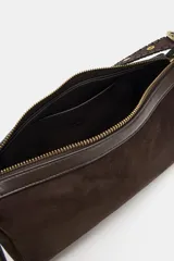 Bolso de hombro de diseño estructurado en color marrón, confeccionado en gamuza. Presenta un asa larga ajustable con detalle de trenzado y hebilla metálica dorada. Cuenta con cierre de cremallera con doble tirador y bolsillo interior.