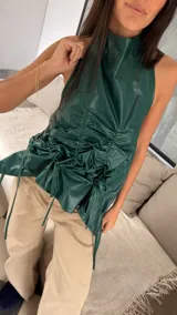 Top de cuero color verde esmeralda con escote halter y espalda descubierta. Presenta un diseño con jaretas frontales que crean un efecto fruncido y ajustable mediante tiras laterales.