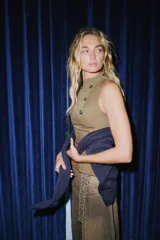 Top sin mangas color verde oliva, con cuello alto y cierre frontal de botones metálicos decorativos. Presenta un corte ajustado en el torso que se ensancha ligeramente hacia el bajo, con detalles de trabillas horizontales a la altura de la cadera.