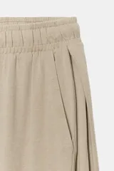 Pantalón de algodón color beige con tiro medio, cintura elástica ajustable con cordón y perneras anchas con pliegues delanteros y bolsillos laterales.