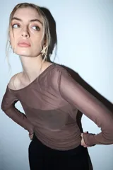 Remera de manga larga, color marrón oscuro, confeccionada en tul transparente con escote barco que cae sobre un hombro. Presenta frunces en el torso y mangas ajustadas.