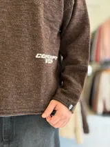 Sweater de punto liviano color beige, con textura marcada, cuello redondo y mangas largas de corte relajado, con pequeño estampado tipográfico negro en el lateral inferior.