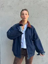 Campera bomber de denim azul oscuro con costuras a contratono en color verde. Presenta cuello de corderito en color marrón, cierre frontal y diseño holgado.