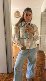 Camisa de manga larga con estampado cuadrillé en tonos beige y blanco, con cuello camisero y bolsillo frontal adornados con volados en color turquesa.