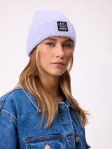 Gorro tipo beanie de punto acanalado en color lila, con parche rectangular negro en el frente que incluye la inscripción 'EAT THE WAVE'.