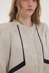 Camisa de lino color beige, de silueta romántica con escote a la base y mini volado. Cuenta con mangas 3/4 abullonadas con frunces y terminación en elástico. Posee detalles de puntillas negras en contraste en el frente y cierre frontal con botones ocultos bajo cartera.