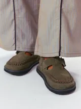 Slipper tipo guillermina confeccionada en gamuza color marrón, con plataforma de goma negra y doble hebilla ajustable. Presenta un pespunte decorativo en el borde inferior y cuenta con interior recubierto en cuero.