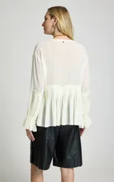 Camisa fluida color blanco marfil con escote en V y cordón ajustable. Cuenta con mangas largas anchas, puños con pliegues y volantes, y dobladillo redondeado.