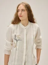 Camisa de lino color crudo, con cuello mao y cierre frontal con botones. Cuenta con detalles de alforzas verticales y diseño bordado floral en el frente, además de mangas 3/4 con volumen y puños elásticos.