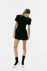 Vestido mini negro con cuello redondo, manga corta con pliegues y costuras marcadas. Cuenta con cierre posterior con cremallera oculta.