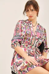 Vestido corto de cuello redondo con escote en V, mangas 3/4 con puños elásticos y lazo ajustable en la cintura. Presenta un estampado multicolor con motivos geométricos y florales.