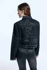 Campera de denim negro con corte estructurado a la cintura, decorada con patrones de tachas metálicas en hombros, mangas y frente. Presenta cierre frontal con botones, bolsillos con solapa y puños abotonados.