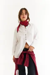 Camisa blanca de algodón con corte oversize, cuello clásico, cierre frontal con botones en contraste y aberturas laterales. Presenta un diseño con recorte diagonal en el frente y detalle de bordado en zigzag en la espalda.