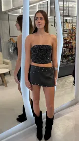 Minifalda de tiro bajo color negro, confeccionada en eco cuero, con diseño cruzado en el frente y detalle de tachas metálicas plateadas a lo largo de los bordes.