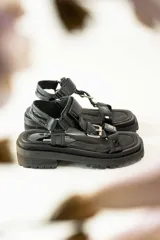 Sandalias negras de estilo chunky con plataforma dentada. Presentan tiras ajustables con cierre de velcro y hebillas metálicas, combinando texturas de cuero granulado y detalles en charol.