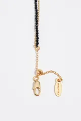 Collar doble dorado, compuesto por una tira corta de cuentas negras con un dije de perla y una cadena más larga con un dije de corazón esmaltado en negro.