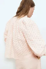 Blusa corta de algodón con diseño broderie de color rosado claro, cuello alto, mangas abullonadas con puños ajustados y cierre frontal de botones con efecto carey.