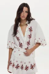 Blusa blanca de gasa de algodón con mangas 3/4 abullonadas y escote en V con botones. Cuenta con bordados en tonos marrones y naranjas en la zona frontal y las mangas.