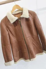 Campera de cuero napalán color marrón, de corte recto con cierre frontal de cremallera, cuello y puños de corderito sintético blanco, bolsillos laterales con ribete y diseño de paneles pespunteados.