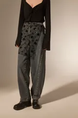 Falda mini de tiro alto confeccionada en denim gris oscuro con efecto lavado. Presenta un diseño superpuesto de tul negro con bordados florales y lentejuelas, además de detalles de tachas metálicas a lo largo de las costuras laterales.