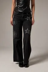 Pantalón jean de corte recto y tiro bajo, color negro gastado, con detalles de apliques de strass formando diseños de estrellas, soles y flores.