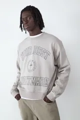Buzo de cuello redondo y manga larga en color gris claro. Presenta un estampado frontal con tipografía estilo universitario y un diseño gráfico central. Cuenta con puños y cintura acanalados.