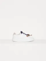 Zapatillas urbanas de cuero vacuno color blanco, con interior al tono y talonera en descarne color beige. Presentan doble cordón, uno azul y otro marrón, y dijes colgantes.