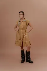 Vestido corto beige de algodón con cuello camisero y cierre frontal de botones. Presenta mangas cortas y detalles bordados a contratono en la zona del pecho y en los bolsillos, además de un volado en el ruedo.