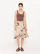 Pollera midi de gabardina color beige con estampado de flores y bordados. Presenta un cruce diagonal con botón dorado personalizado y botón interno para sostener la solapa inferior. Incluye bolsillo plaqué en delantero orientado a cintura y volado en forma de caracol en el ruedo.