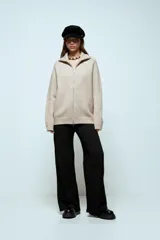 Cardigan de punto oversize en color beige, con cuello alto y cierre frontal completo de doble cursor. Presenta mangas largas con puños acanalados y un diseño holgado.