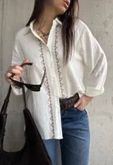 Camisa blanca de tejido liviano y calce relajado, con manga larga y detalle de puntilla y bordado en contraste a lo largo de la botonadura frontal.