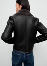 Chaqueta estilo biker de efecto piel en color negro, con cuello y forro interior de pelo sintético en tono gris oscuro. Presenta cierre frontal de cremallera, manga larga y diseño estructurado.