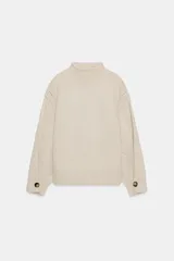 Jersey de punto color beige con cuello subido, manga larga y detalle de botones en los puños. Presenta costuras marcadas y acabados en rib.