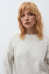 Sweater de punto en color beige melange, con cuello redondo y mangas largas. Presenta un diseño decorativo con perlas aplicadas en la parte frontal y en las mangas.