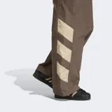 Pantalón deportivo Adidas de corte holgado, color marrón con detalles en beige en la parte inferior de las piernas. Presenta cintura elástica con cordón de ajuste y logo de la marca bordado en el muslo.