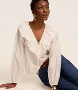 Blusa color blanco confeccionada en voile, de mangas largas con elástico en los puños, cuello en V con volado y lazo con adornos metalizados.