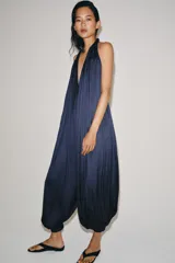 Mono largo satinado de color azul marino, con cuello halter, escote en V profundo y espalda descubierta. Presenta diseño plisado y bajo abullonado con terminación en puño.