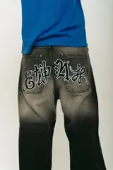 Pantalón de jean negro con lavado localizado (efecto tie-dye o acid wash), corte baggy y amplio. Presenta apliques bordados en la parte trasera de los bolsillos y herrajes metálicos cromados.