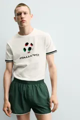 Camiseta de corte relajado confeccionada en piqué de algodón color crudo. Presenta cuello redondo, mangas cortas con ribetes a contraste y estampado frontal con el logo de Italia 90 FIFA World Cup.
