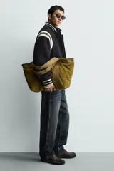 Bolso tipo shopper de gran capacidad, confeccionado en tejido con textura de pana. Cuenta con dos asas de mano resistentes, un bolsillo frontal plano y cierre principal mediante botón a presión. El interior incluye un bolsillo con cierre de cremallera.