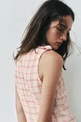 Vestido corto sin mangas de tejido texturizado con diseño de cuadros en tonos crudo y fucsia. Presenta cuello subido, trabillas decorativas con botones dorados en los hombros y cierre lateral con cremallera oculta.