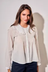 Blusa blanca de algodón peinado con mangas largas, cuello babero bordado y pliegues frontales.