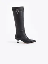 Bota de caña alta confeccionada en cuero vacuno liso, con taco laqueado de altura intermedia y punta fina. Presenta detalles de hebillas metálicas en la caña y en la puntera, incluyendo un dije en forma de corazón.