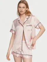 Pijama corta de satín con diseño de rayas verticales rosadas y blancas, ribetes negros en cuello, solapa, bolsillo y dobladillos. Consta de una camisa de manga corta con botones frontales y bolsillo en el pecho, y un short a juego con cintura elástica.
