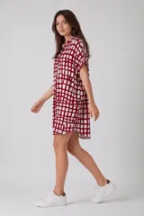 Vestido camisero de corte holgado y silueta relajada, con cuello clásico, cierre frontal de botones y mangas cortas con dobladillo. Presenta un estampado de cuadros irregulares en color bordó sobre fondo blanco.