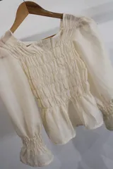 Blusa de viyela color crema con cuerpo fruncido tipo nido de abeja, escote cuadrado y mangas largas abullonadas con puños elásticos.