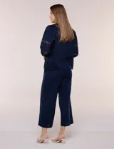 Blusa blanca de calce suelto y caída ligera, con escote en V y cuello Mao pequeño. Presenta mangas tres cuartos abullonadas con elástico en los puños y detalles de calado o puntilla encastrada. Espalda lisa con canesú y leve frunce.